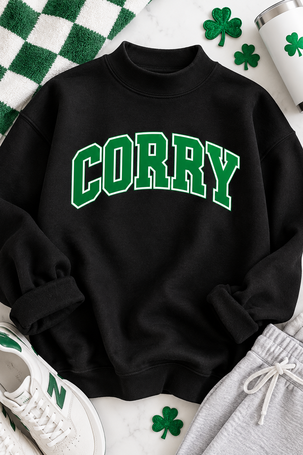 A black crewneck sweatshirt with 'CORRY' printed in green varsity letters across the chest, styled in a flat lay with St. Patrick's Day themed props including shamrocks, a green checkered towel, a white New Balance sneaker, and gray sweatpants.