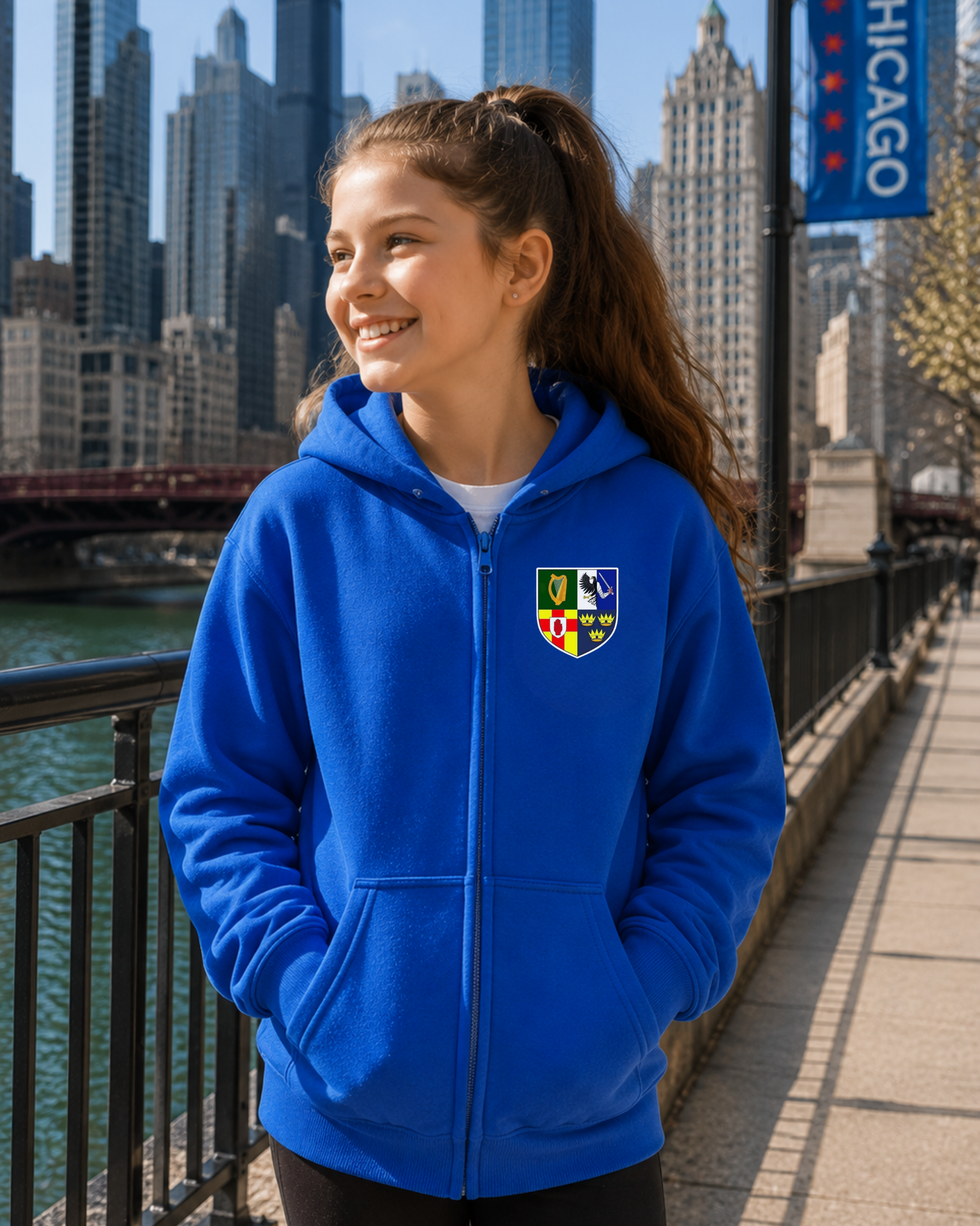 A smiling young woman stands on a riverside walkway wearing a bright royal blue zip-up hoodie with a multicolor crest patch on the left chest. The background shows a city skyline, river, bridge, and a Chicago banner.