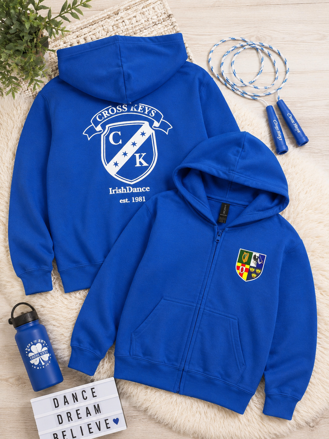 A royal blue zip-up hoodie shown from both front and back in a styled flat-lay. The front view shows a small multicolor shield crest on the left chest, and the back view shows a larger white crest with the text 'Cross Keys IrishDance est.