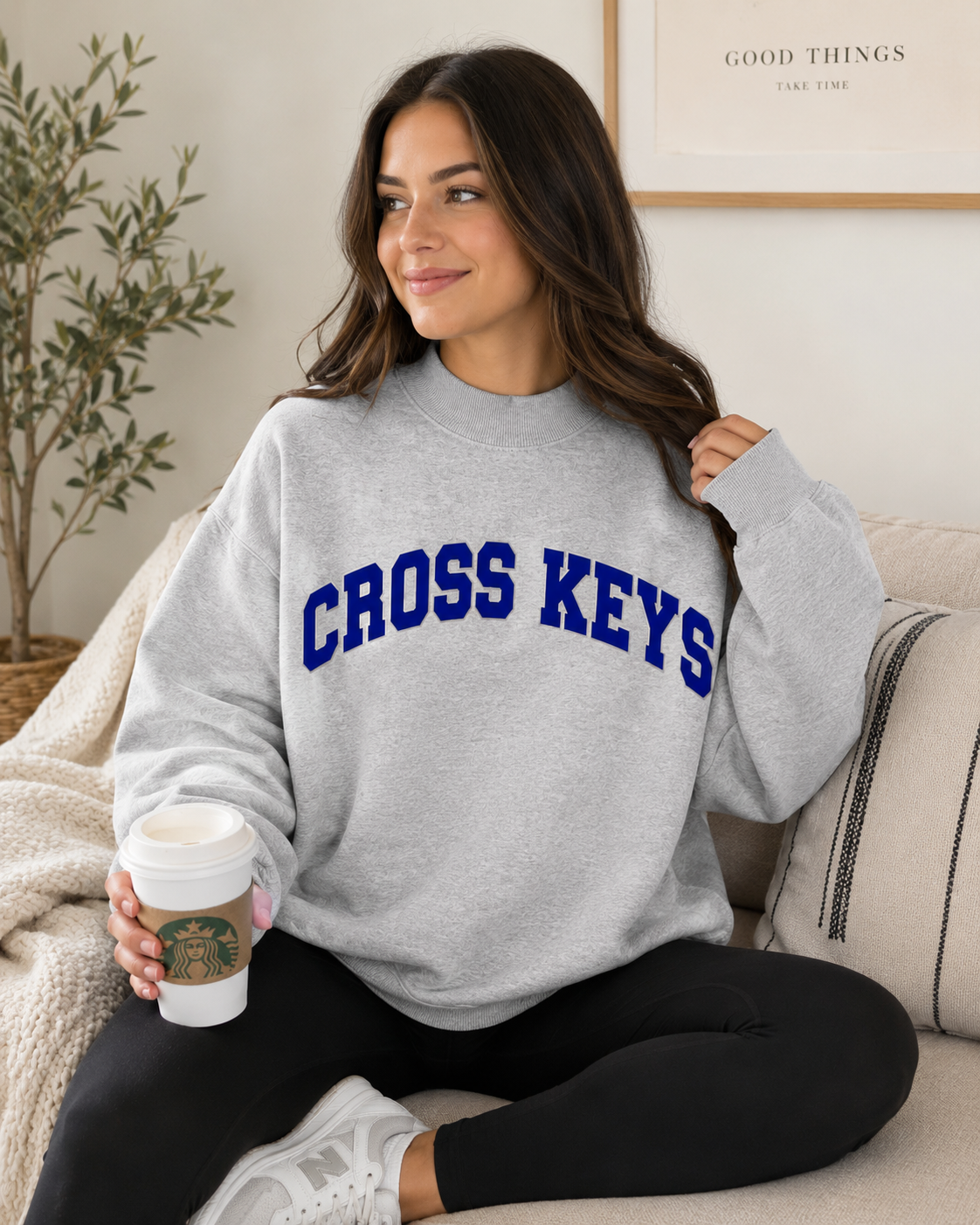 A woman sitting on a beige couch wearing a light gray crewneck sweatshirt with 'CROSS KEYS' printed in blue arched letters across the chest. She is holding a white Starbucks coffee cup.