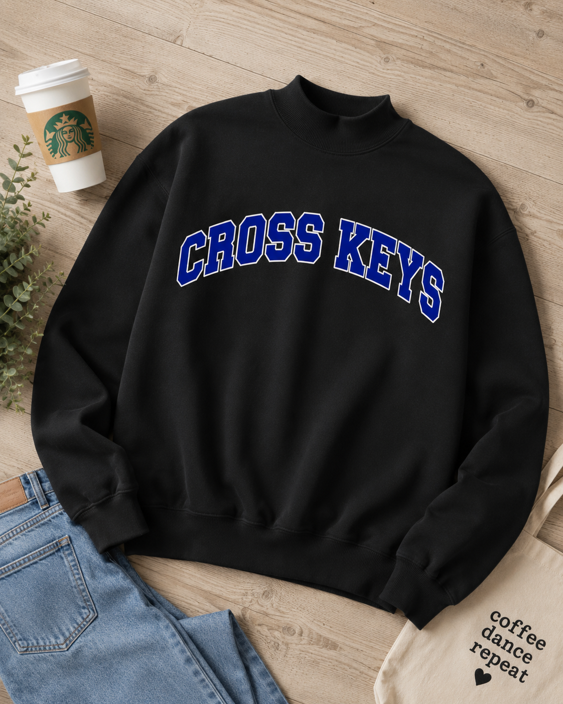 A black mock-neck sweatshirt with 'CROSS KEYS' printed in blue varsity-style lettering across the chest, styled in a flat lay on a light wood floor with jeans, a Starbucks cup, and a tote bag nearby.