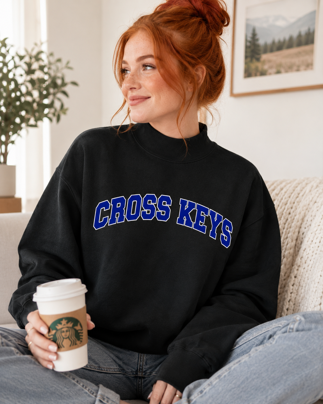 A woman with red hair in a bun sits on a couch holding a Starbucks coffee cup, wearing a black sweatshirt with 'CROSS KEYS' printed in blue and white arched lettering across the chest.