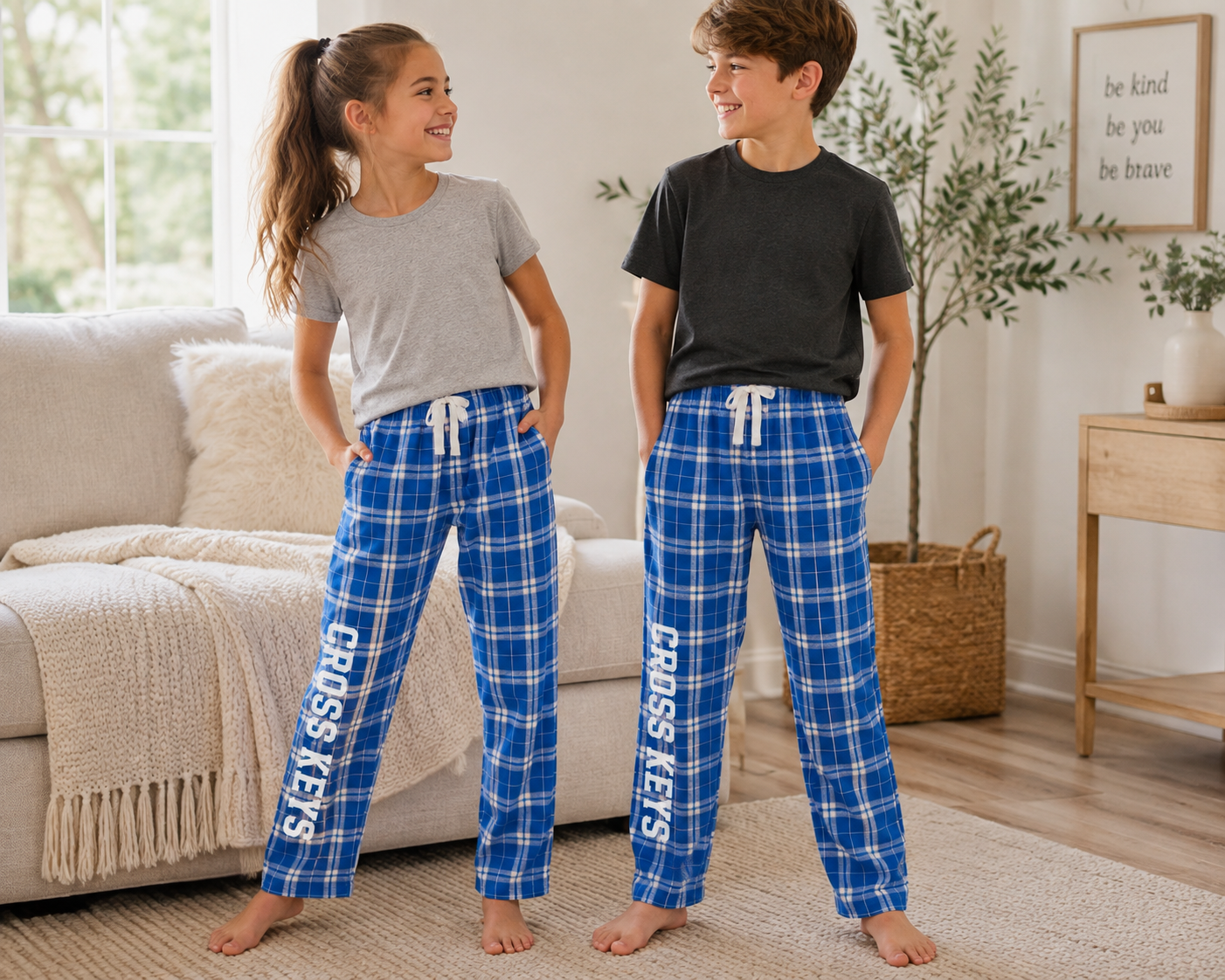 A boy and a girl standing in a bright living room, both wearing matching blue plaid pajama pants with white drawstrings and vertical 'CROSS KEYS' text down the left leg. The girl also wears a light gray t-shirt, and the boy wears a dark charcoal t-shirt.