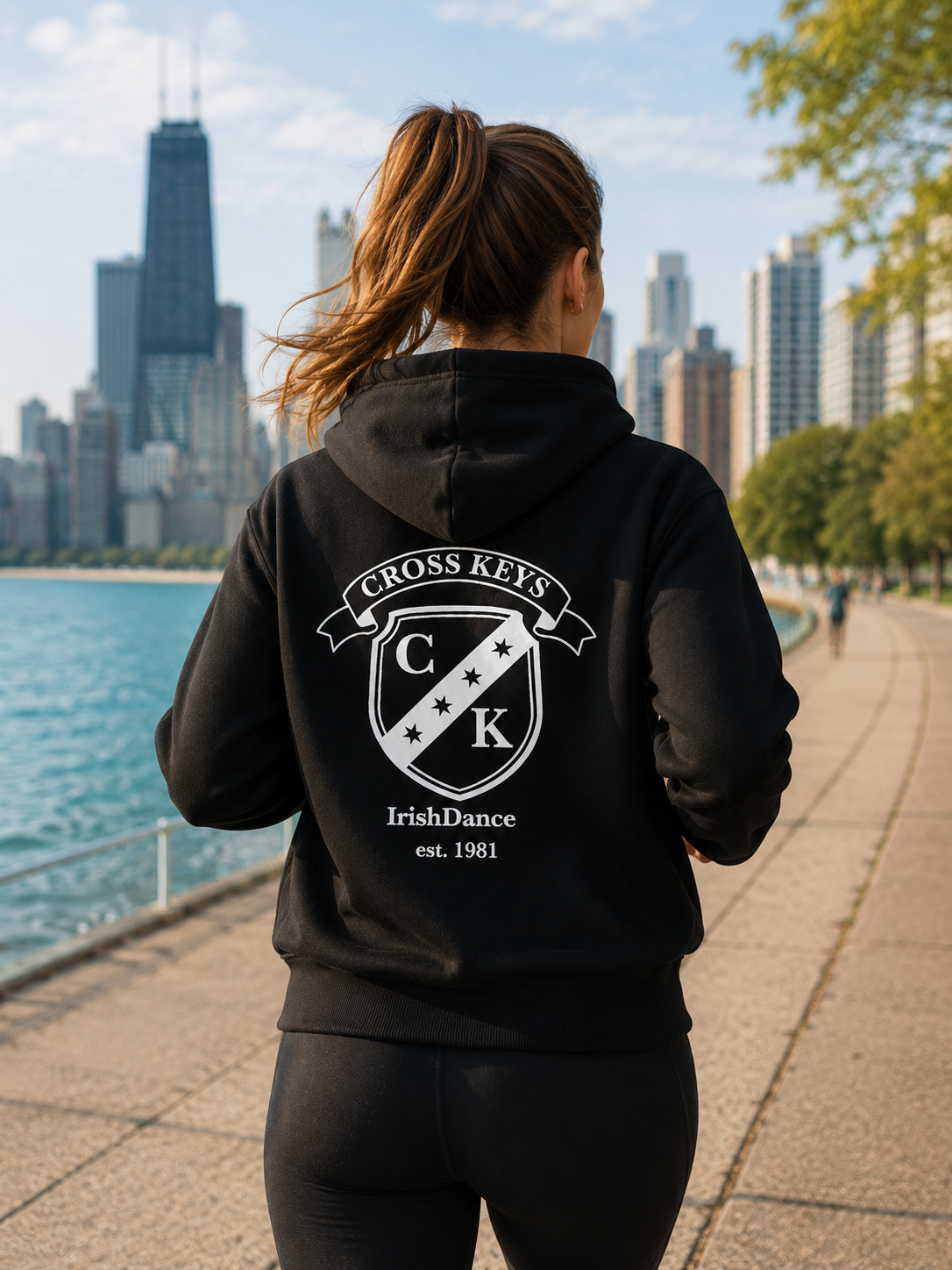 A woman viewed from behind wearing a black hoodie with a large white 'Cross Keys IrishDance' graphic on the back, standing on a waterfront path with a city skyline in the background.