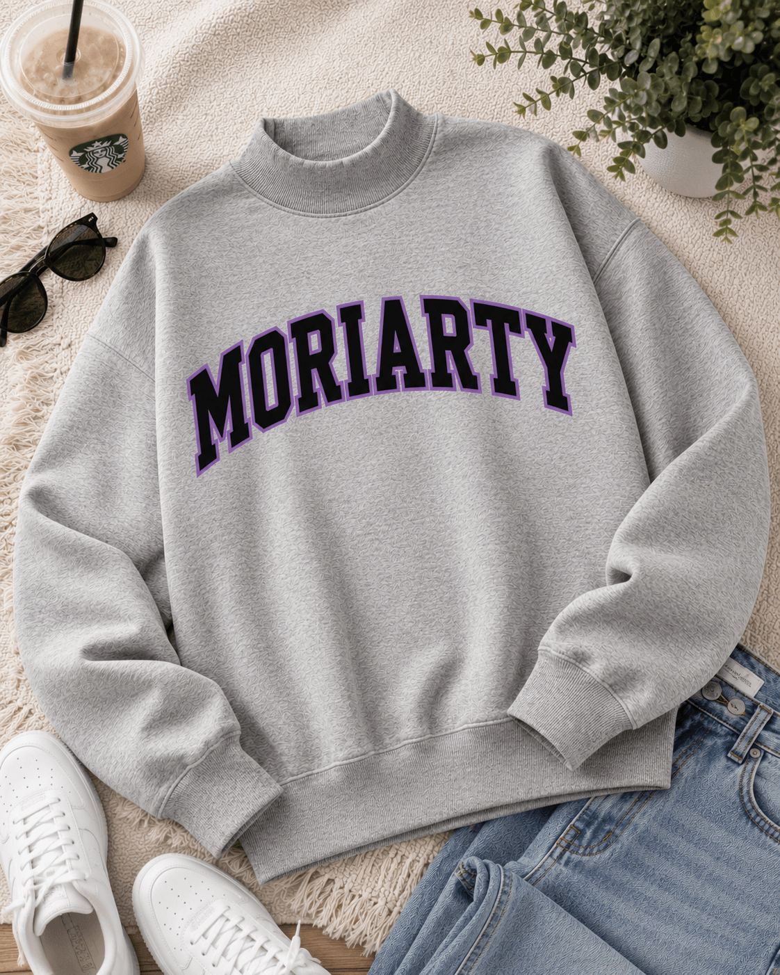 A light gray crewneck sweatshirt with 'MORIARTY' printed across the chest in dark purple varsity lettering, styled in a flat lay with jeans, white sneakers, sunglasses, an iced coffee, and a potted plant.