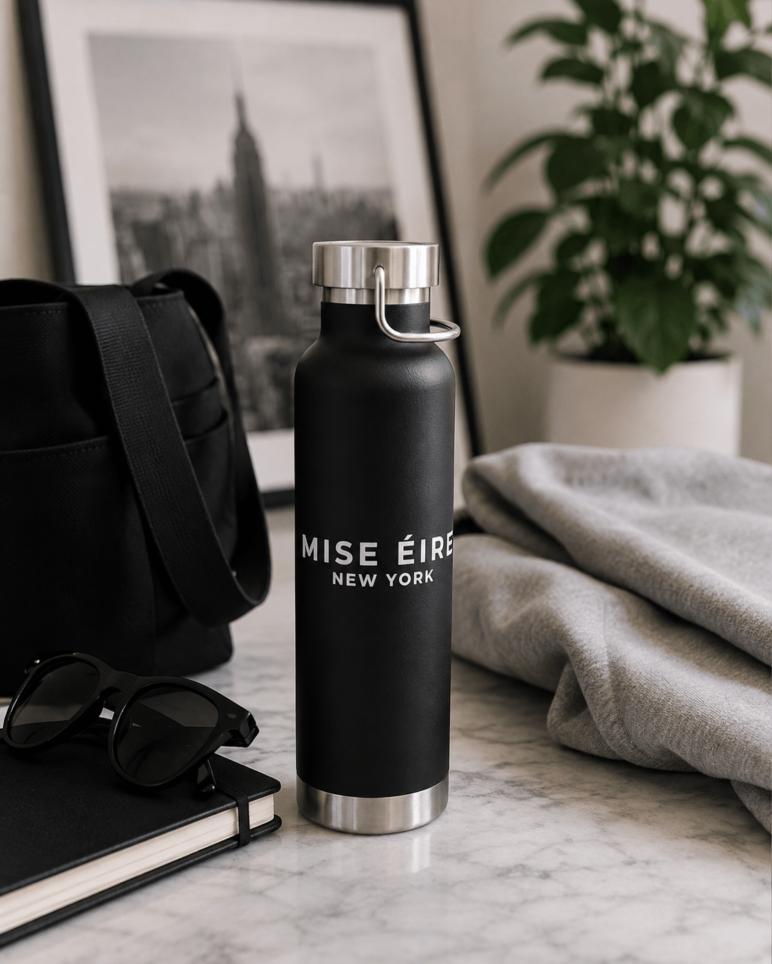 A matte black insulated water bottle with silver metal accents stands on a marble surface in a styled indoor setting. The bottle has the text 'MISE ÉTIQUE NEW YORK' printed on the front.