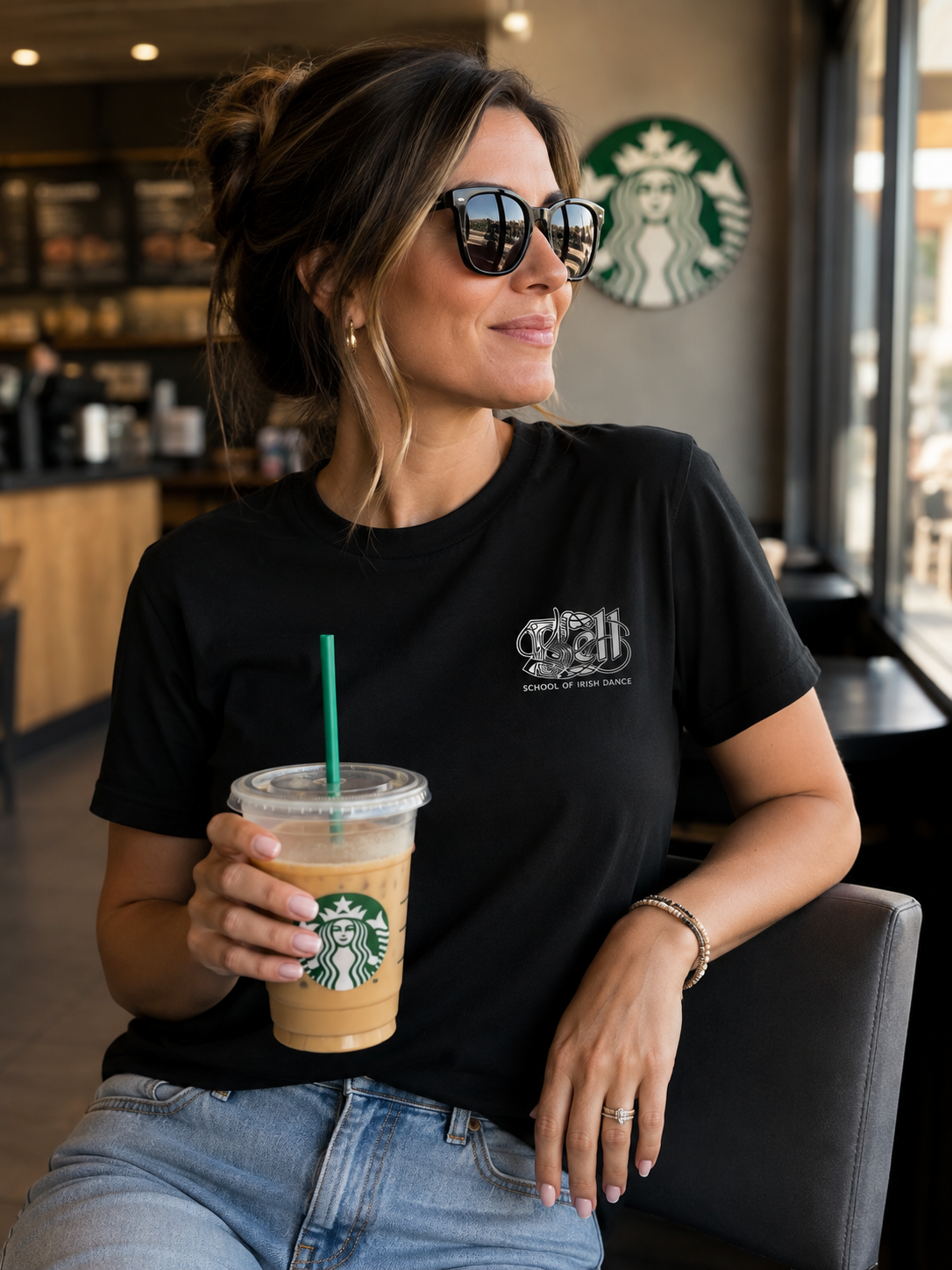 A woman wearing a black graphic t-shirt and sunglasses sits in a Starbucks cafe holding an iced coffee. The t-shirt features a small white crest logo on the left chest with the text 'Crown of My Own'.