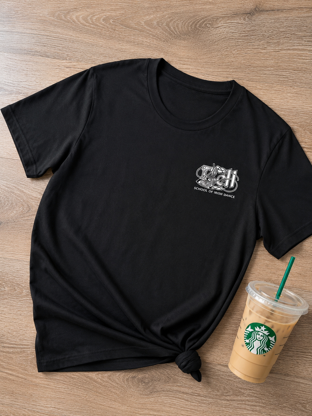 A black short-sleeve T-shirt laid flat on a wood surface, with a small white chest graphic on the wearer-left side and a tied hem. A Starbucks iced coffee cup with a green straw sits to the lower right as a styling prop.