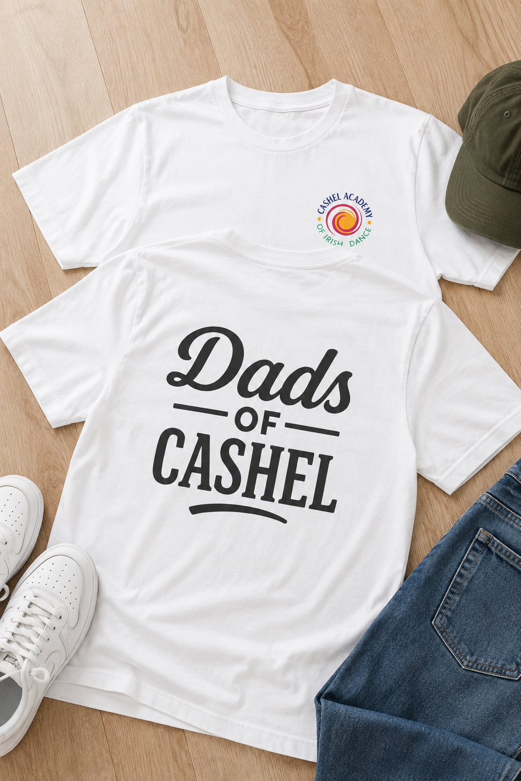 A white graphic T-shirt laid flat on a light wood floor, with the words 'Dads of Cashel' printed in large black lettering on the front. A small circular logo appears on the upper left chest.