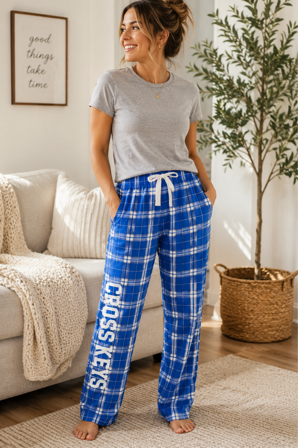 A woman stands barefoot in a bright living room wearing blue plaid pajama pants with a white drawstring and large white 'CROSS KEYS' text down one leg, paired with a gray t-shirt.