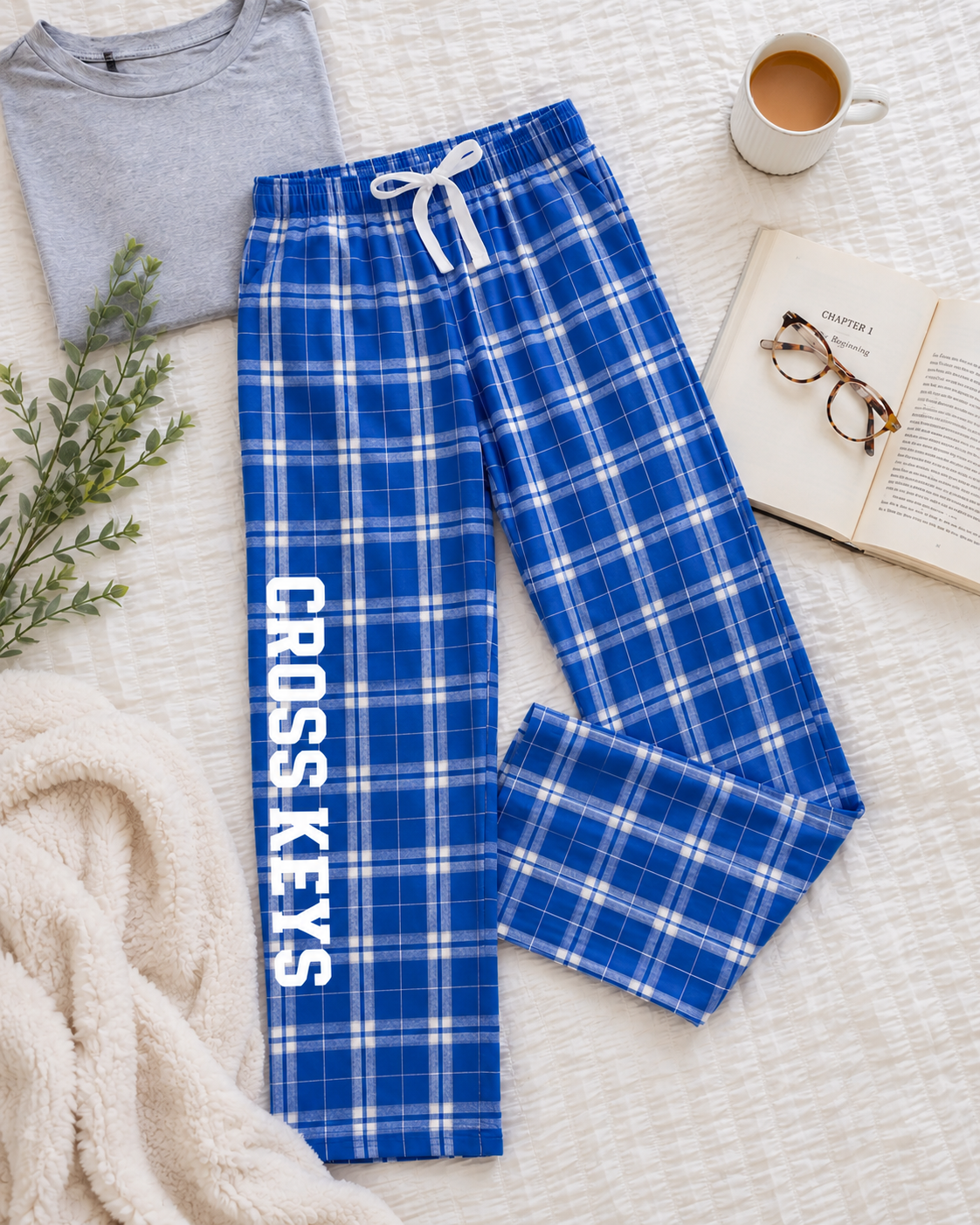 Blue plaid pajama pants with white stripes and 'CROSS KEYS' printed vertically in white on one leg, styled in a cozy flat lay with a gray t-shirt, mug, book, glasses, and blanket.