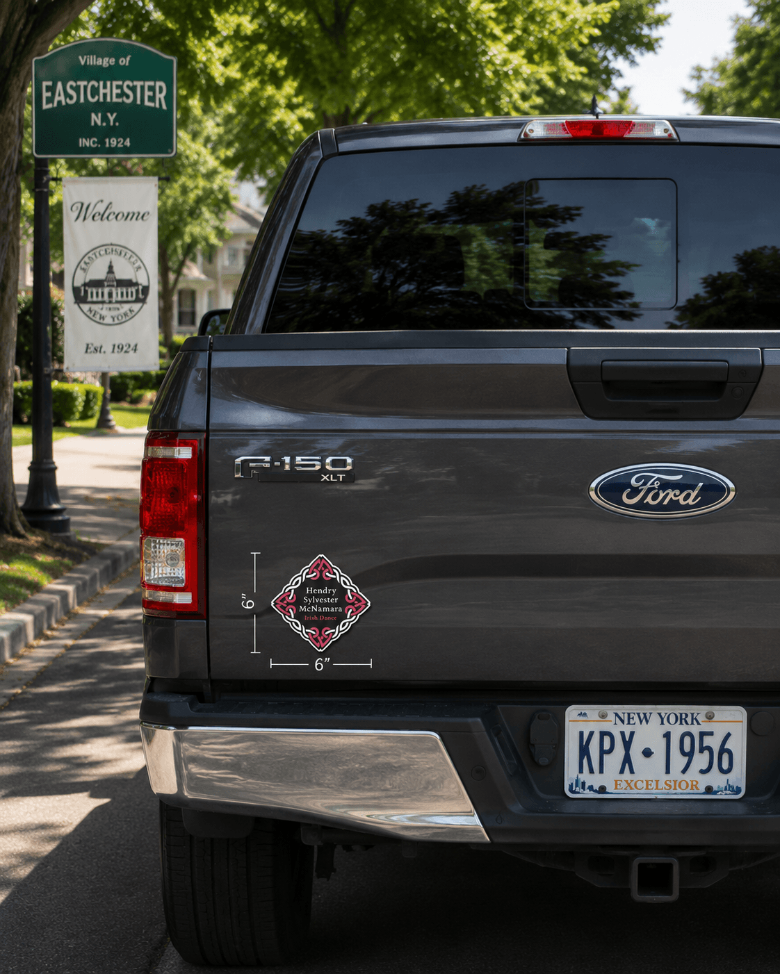 A dark gray Ford R-150 pickup truck shown from the rear, parked outdoors on a street with trees and a Village of Eastchester welcome sign in the background. A small diamond-shaped decorative decal is visible on the left side of the tailgate.