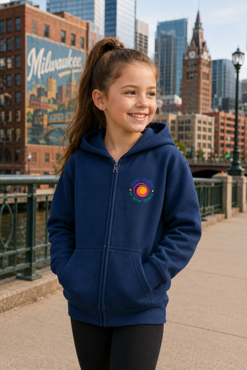 A young girl smiling and looking to the side while wearing a navy blue zip-up hoodie with a colorful circular logo on the left chest. The background shows a city skyline with brick buildings and a bridge railing.