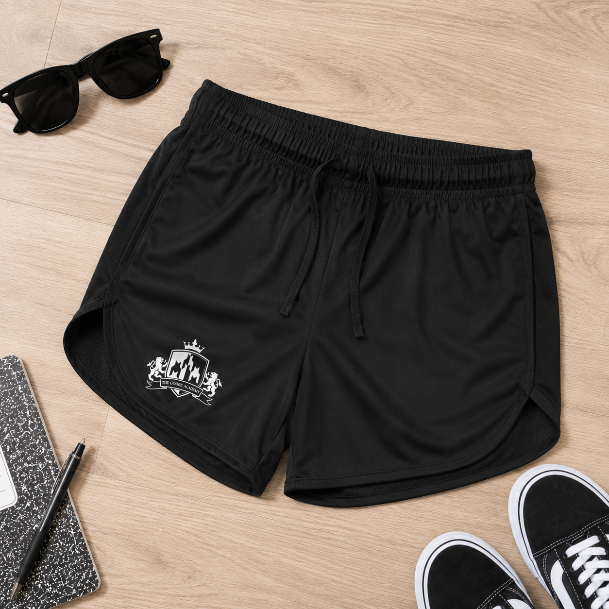 Black athletic shorts with a white graphic print on the left leg, styled in a flat lay on a light wood floor with sunglasses, a notebook, pen, and black sneakers.