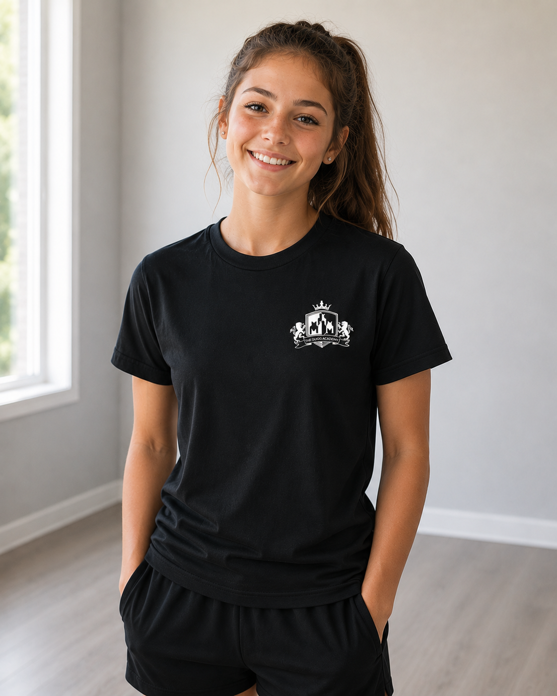A smiling young woman with her hair in a ponytail stands indoors wearing a black short-sleeve crewneck t-shirt with a small white crest graphic on the left chest, paired with black shorts.