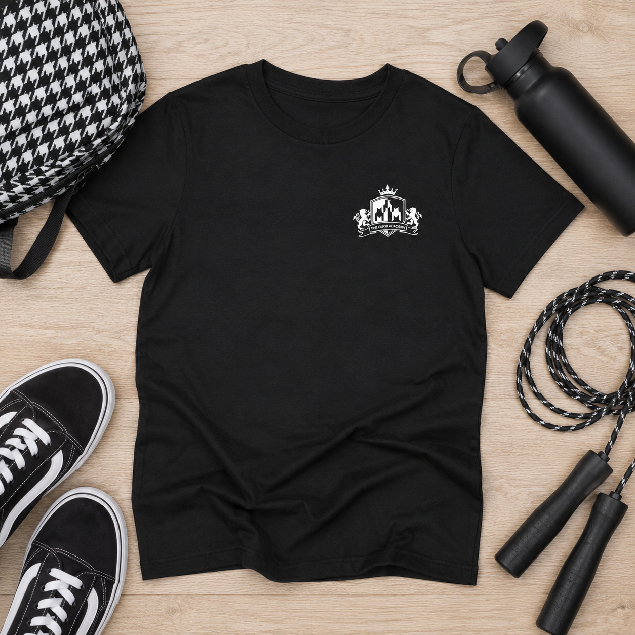 A black short-sleeve crewneck t-shirt with a small white crest graphic on the left chest, presented in a flat-lay arrangement on a light wood background alongside a black-and-white houndstooth backpack, black sneakers, a black water bottle, and a jump rope.