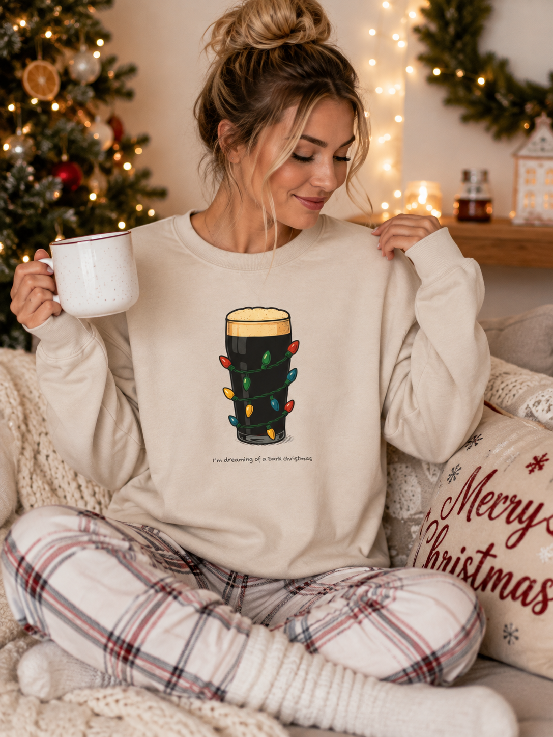 A woman sitting on a cozy couch in a living room decorated for Christmas, wearing a cream-colored sweatshirt with a graphic of a mug wrapped in holiday lights and the text 'I'm dreaming of a holiday singalong'. She is holding a white mug and wearing plaid pajama pants.