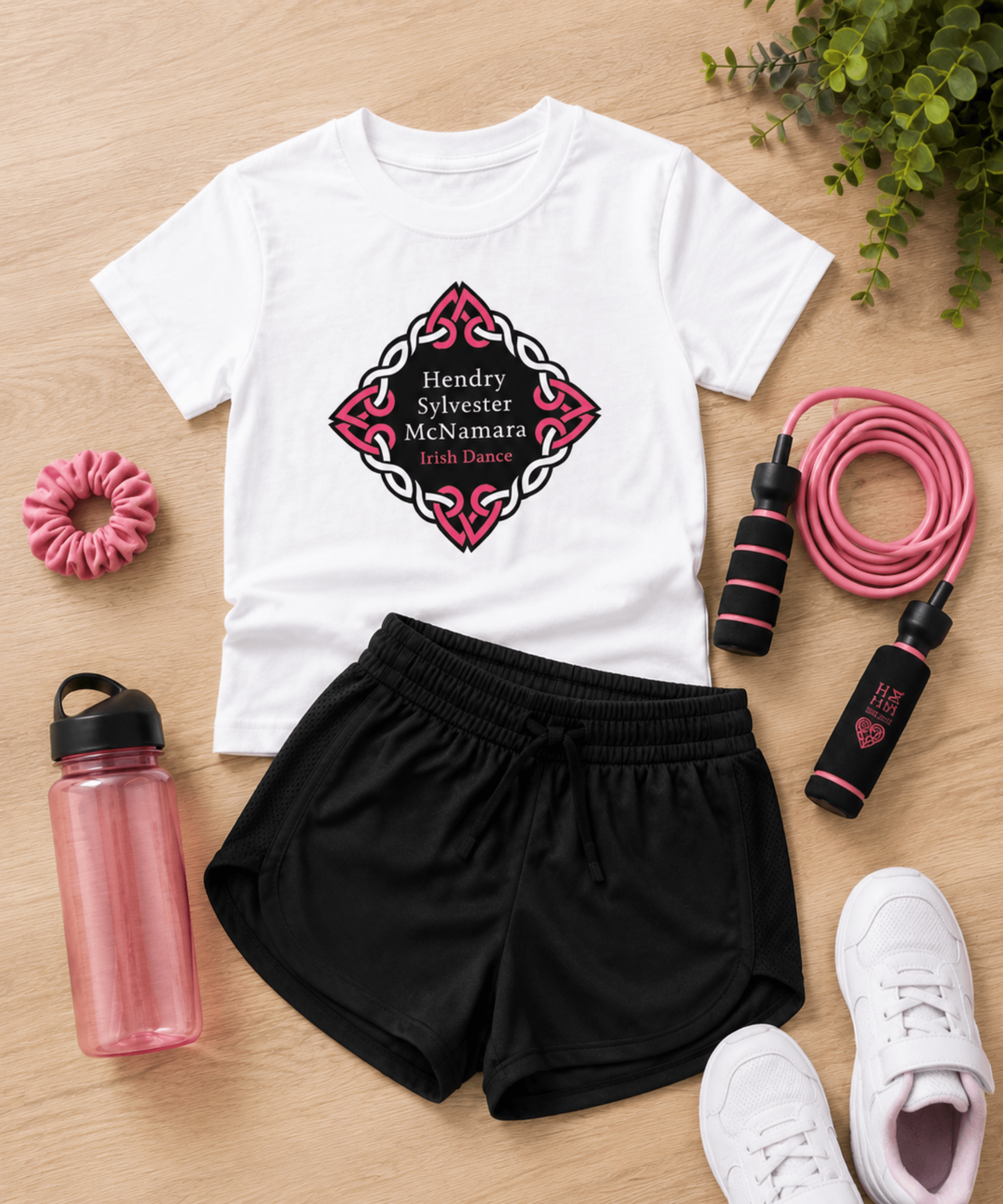 A styled flat-lay of a white graphic T-shirt with black athletic shorts, surrounded by pink and white fitness accessories on a light wood background. The shirt features a Celtic knot diamond graphic with the text 'Hendry Sylvester McNamara Irish Dance'.