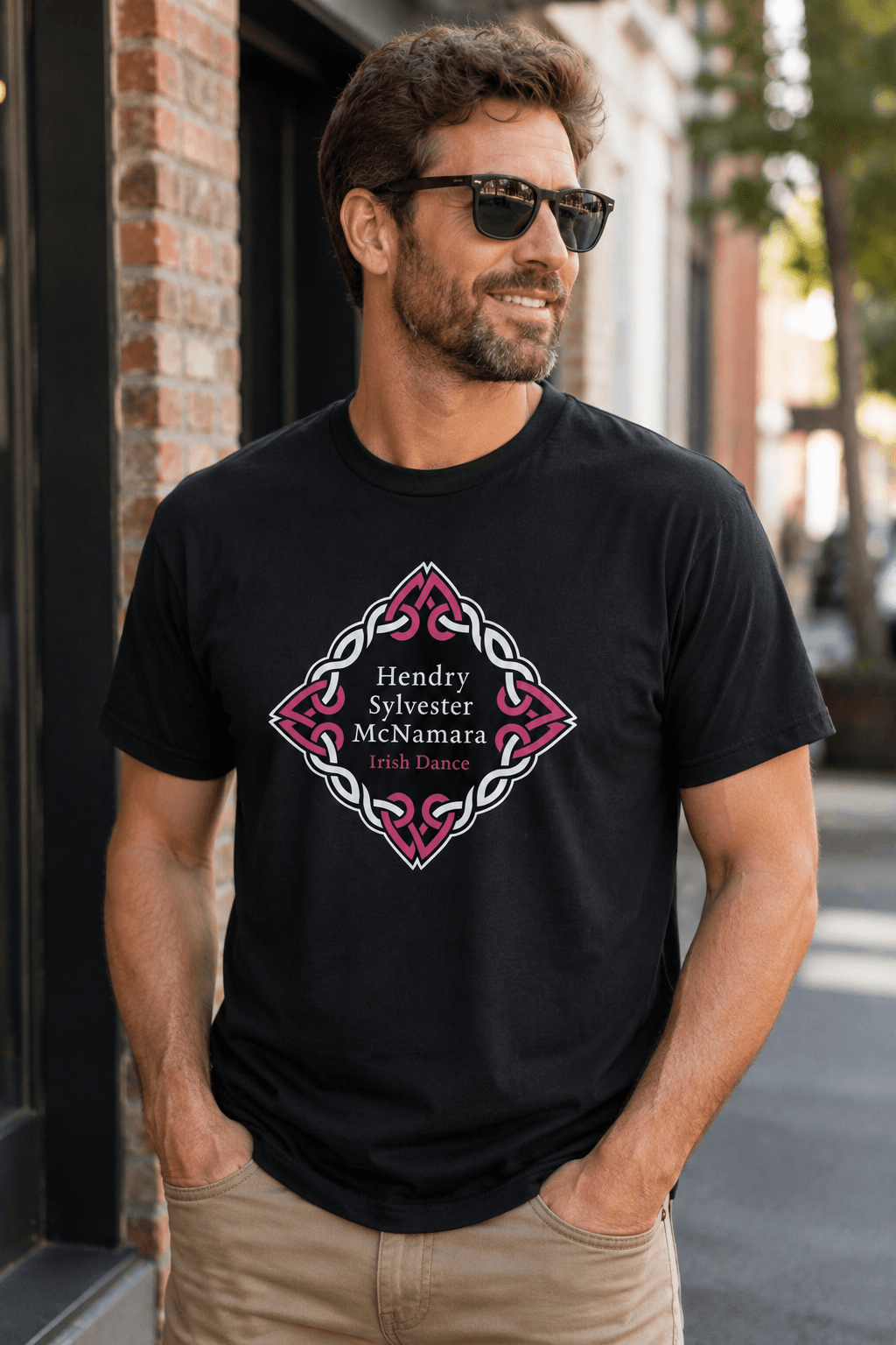 A man wearing a black graphic t-shirt with a pink and white Celtic knot design and the text 'Hendry Sylvester McNamara Irish Dancer' on the front, standing outdoors on a city street.