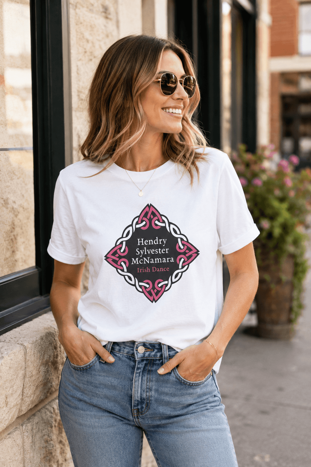 A woman wearing a white graphic t-shirt with a pink and black Celtic knot design and the text 'Hendry Sylvester McNamara Irish Dance', paired with blue jeans, standing outdoors in front of a building.