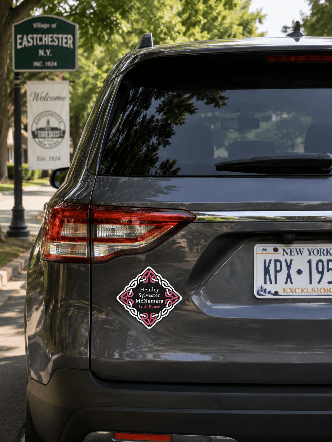 A dark gray SUV rear view on a street with a green Eastchester sign in the background. The vehicle has a decorative diamond-shaped decal on the left side of the rear bumper with red, white, and black text.