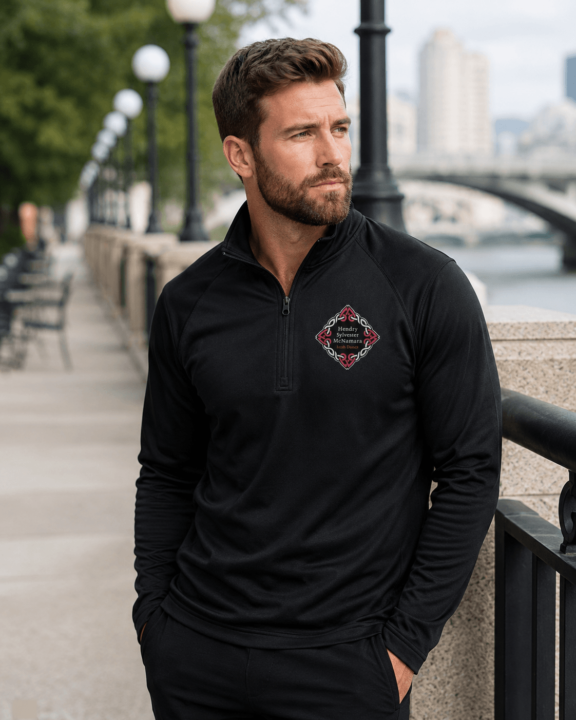 A man wearing a black long-sleeve quarter-zip pullover with a diamond-shaped embroidered logo on the left chest, standing outdoors on a riverside walkway.