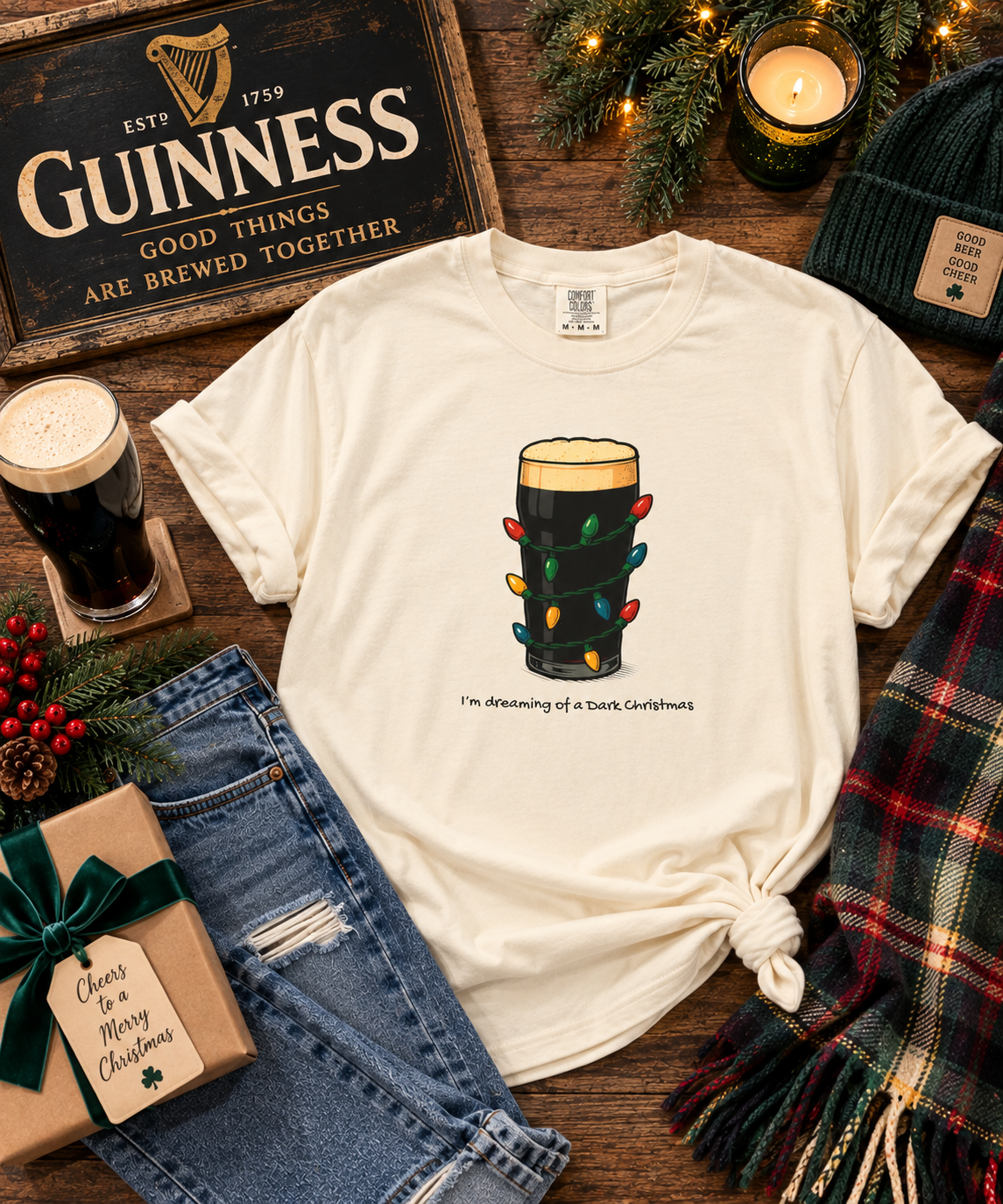 A cream-colored graphic t-shirt styled in a festive flat-lay arrangement on a rustic wooden table. The t-shirt features a graphic of a pint of dark beer decorated with Christmas lights and the text 'I'm meaning of a tank, Carhartt'.