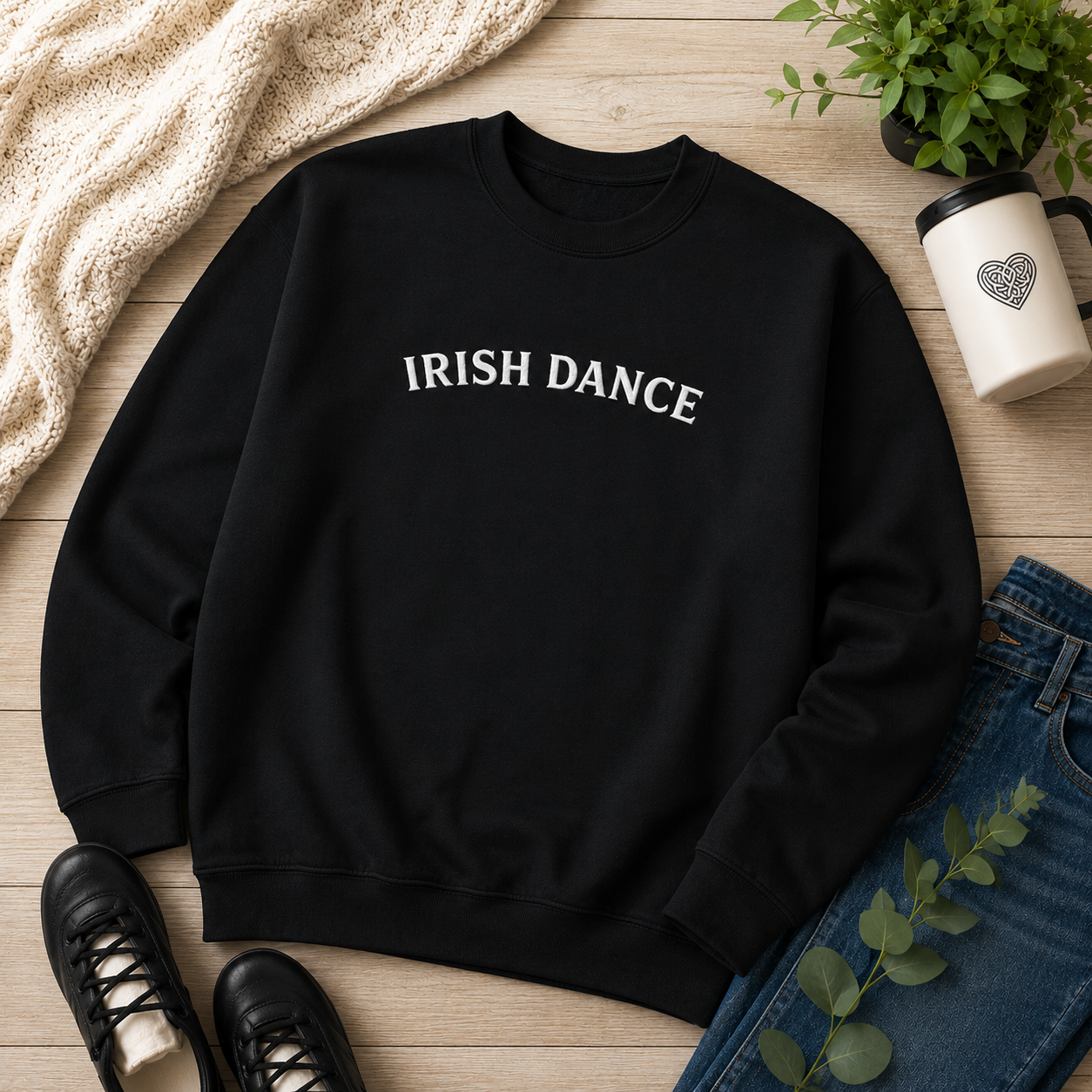 A black crewneck sweatshirt with 'IRISH DANCE' printed in white across the chest, styled in a flat lay on a light wood surface with jeans, black shoes, a cream knit blanket, a plant, and a white travel mug.