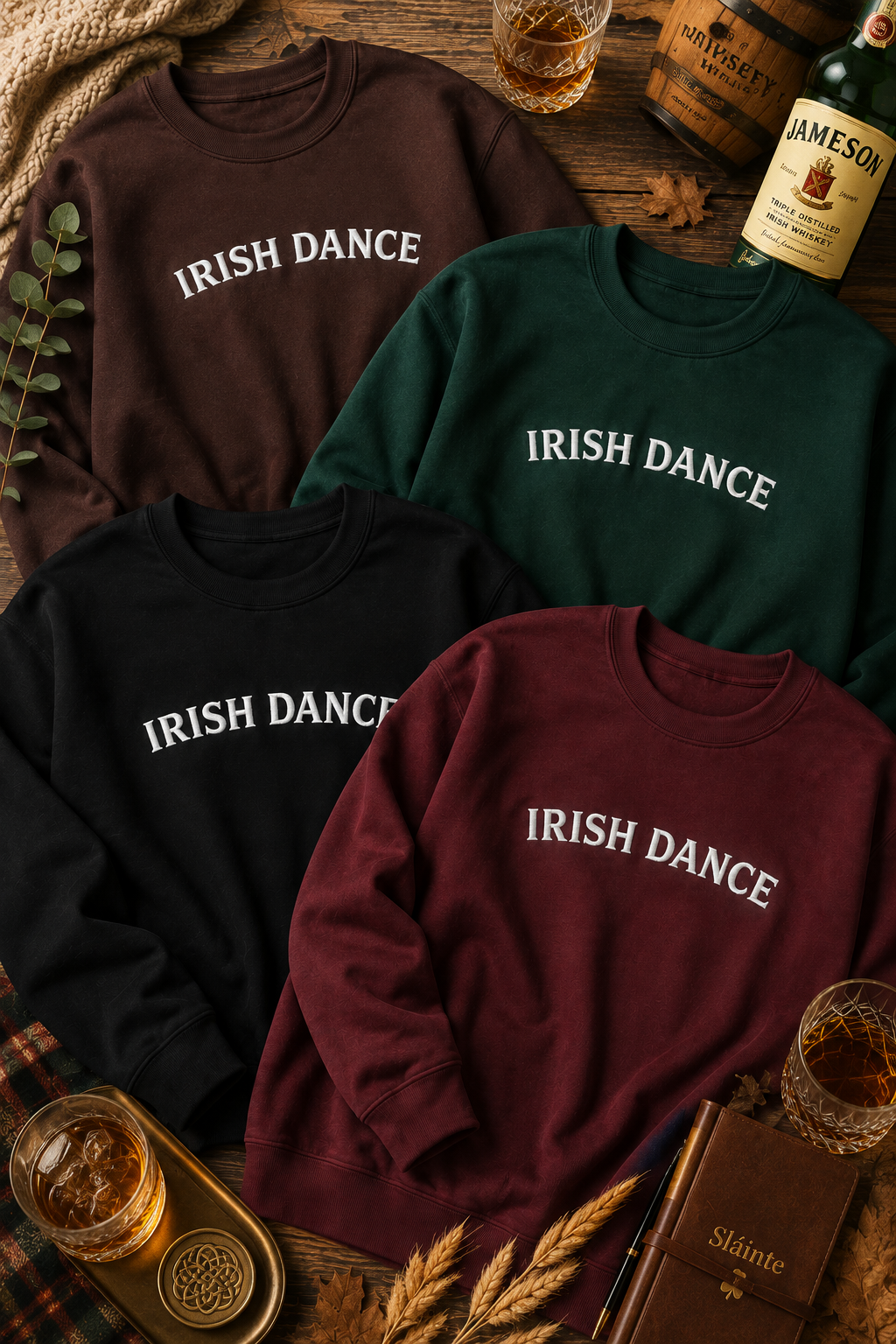 Four crewneck sweatshirts in brown, green, black, and burgundy, each printed with 'IRISH DANCE' in white across the chest. The sweatshirts are arranged in a flat-lay on a rustic wooden table surrounded by whiskey glasses, a Jameson bottle, wheat ears, and a book.