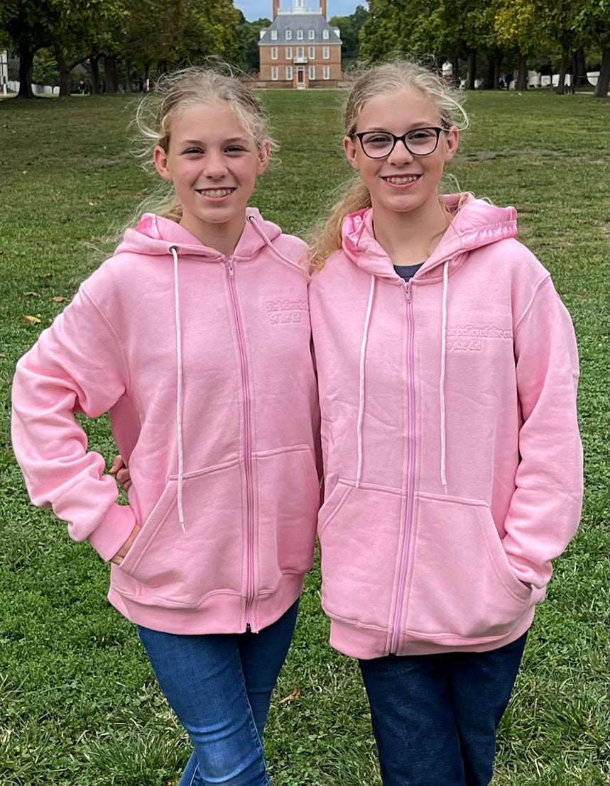 Two girls wearing pink hoodies standing on grass with a building in the background