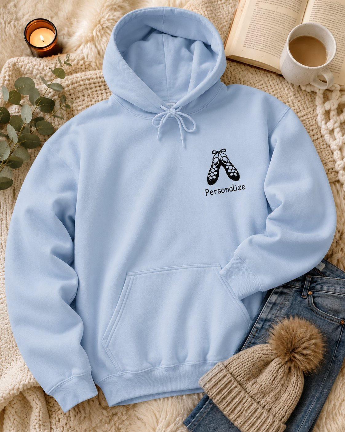 A light blue pullover hoodie with a small black graphic of dancing shoes and the word 'Personalize' on the left chest, styled in a cozy flat-lay with a knit blanket, jeans, a beanie, an open book, a mug of coffee, and a candle.