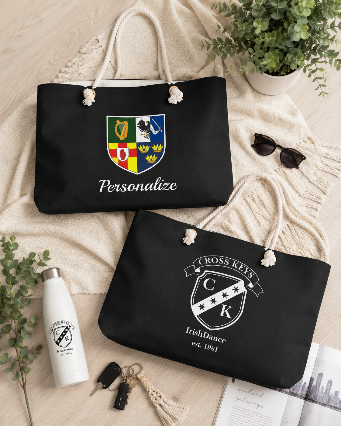 Two black tote bags with cream rope handles are arranged on a beige blanket with lifestyle props. The top bag has a colorful shield crest and the word 'Personalize,' while the bottom bag has a white crest reading 'Cross Keys IrishDance est.