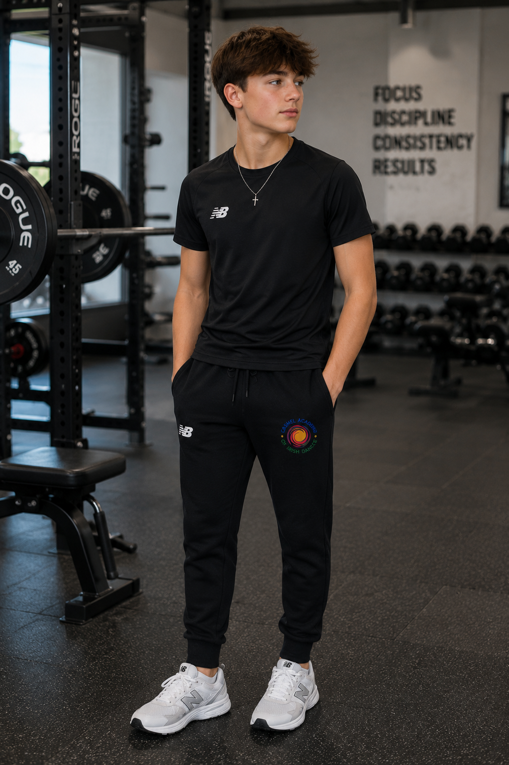 A young man standing in a gym wearing a black New Balance t-shirt and matching black joggers with a circular multicolor graphic on the left thigh.