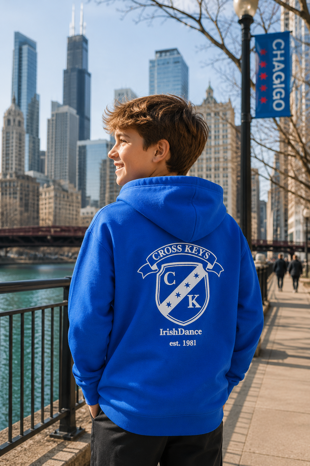 A boy wearing a bright blue hoodie with a white 'Cross Keys Irish Dance' crest graphic on the back, standing on a city sidewalk with a Chicago skyline and river in the background.