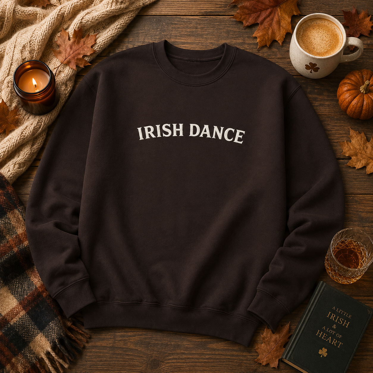 A dark brown crewneck sweatshirt with 'IRISH DANCE' printed in white across the chest, styled in a cozy autumn flat-lay on a wooden table with a mug, candle, scarf, and other seasonal props.