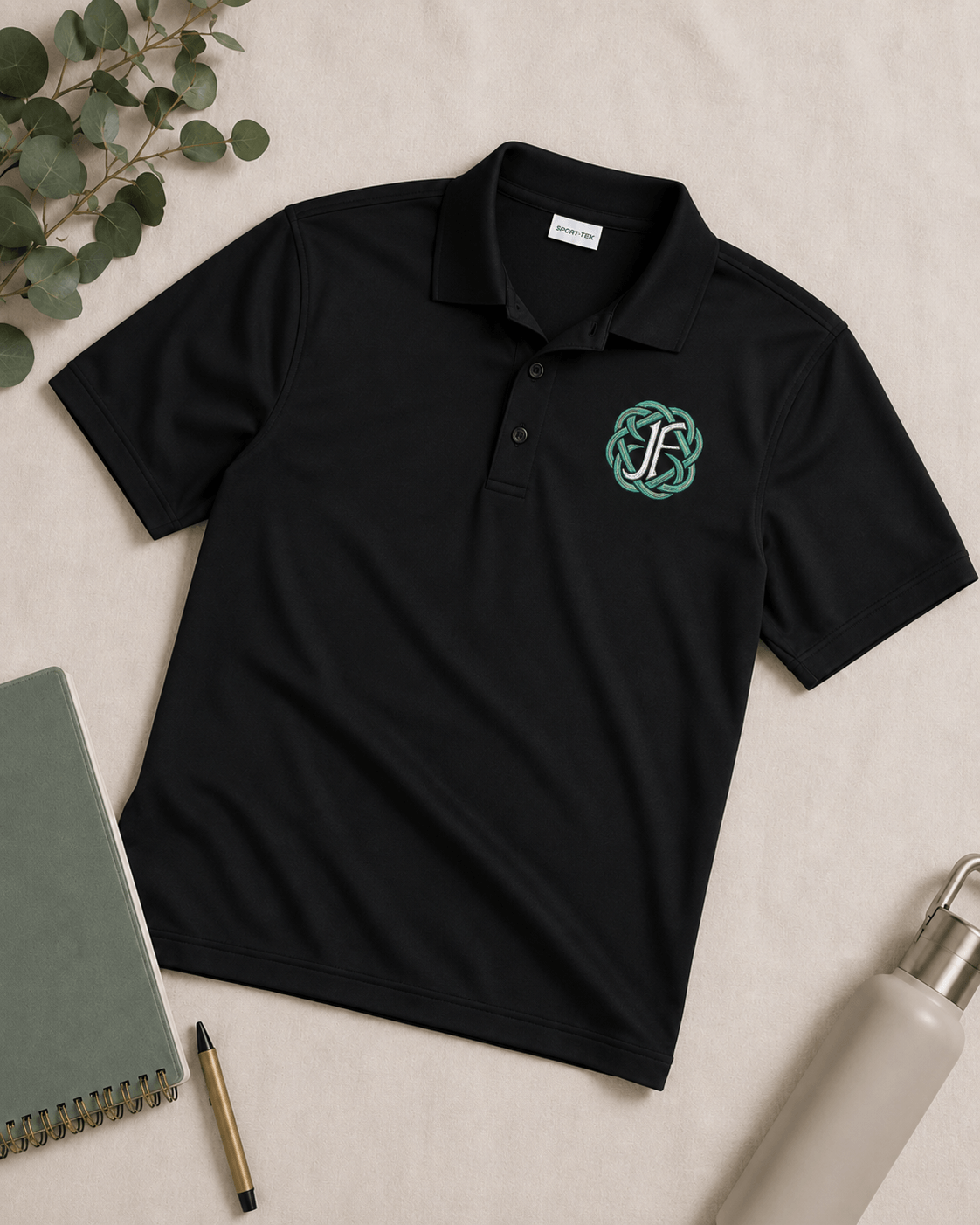 A black short-sleeve polo shirt laid flat on a light beige surface, featuring a green and white Celtic knot monogram embroidered on the left chest. The shirt is styled with a notebook, pen, water bottle, and eucalyptus leaves.