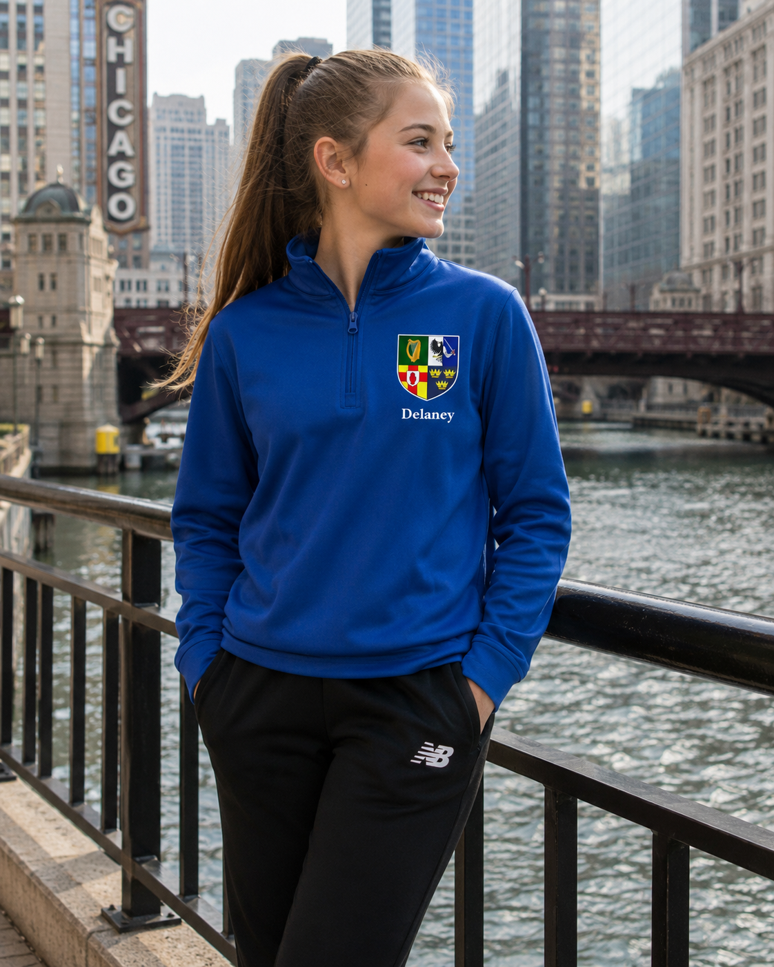 A young woman stands on a bridge over a river with a city skyline in the background, wearing a bright blue quarter-zip sweatshirt with a custom crest and the name 'Delaney' printed on the chest, paired with black sweatpants.