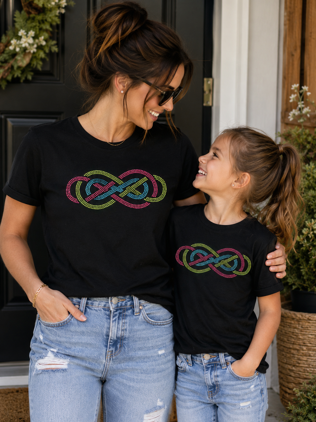 A woman and a young girl wearing matching black graphic t-shirts with a colorful infinity knot design, standing together in front of a home doorway.