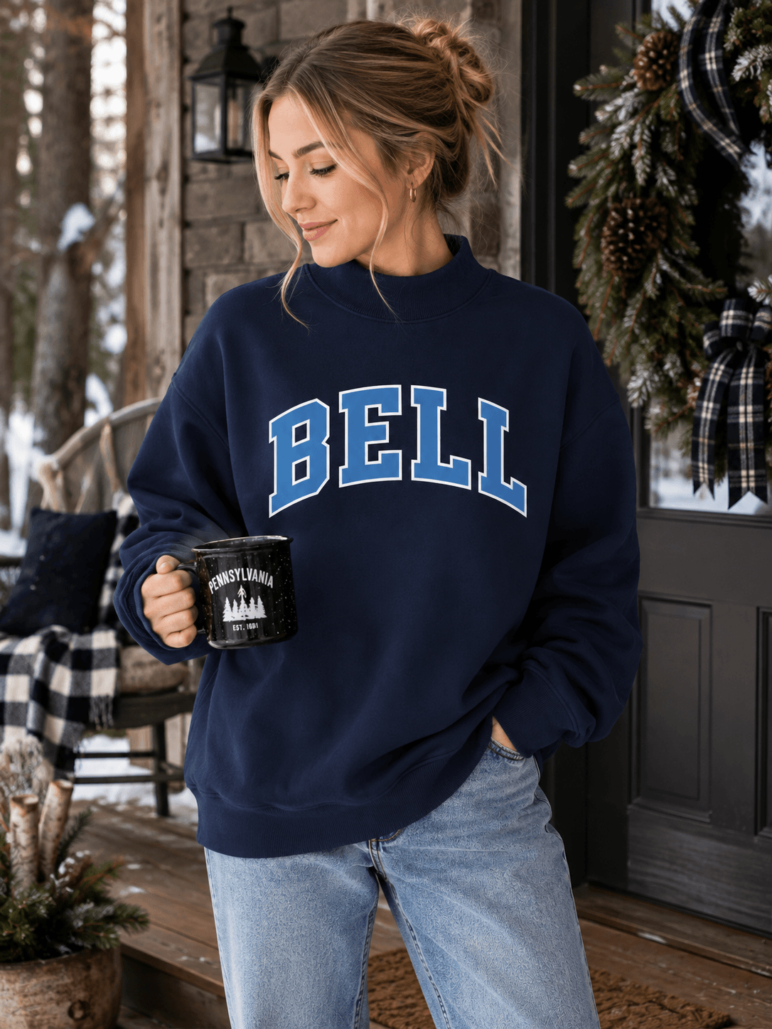 A woman wearing a navy blue sweatshirt with 'BELL' printed in blue varsity letters across the chest, paired with light blue jeans, holding a black mug on a rustic wooden porch.