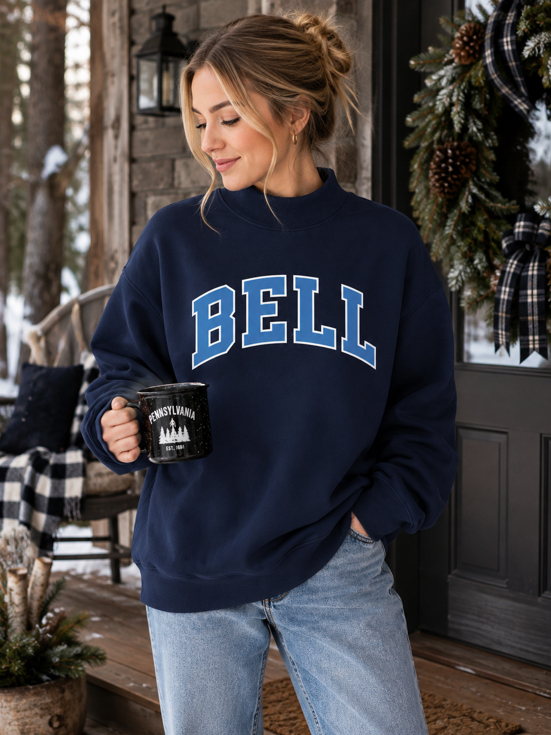 A woman wearing a navy blue sweatshirt with 'BELL' printed in blue varsity letters across the chest, paired with light blue jeans, holding a black mug on a rustic wooden porch.