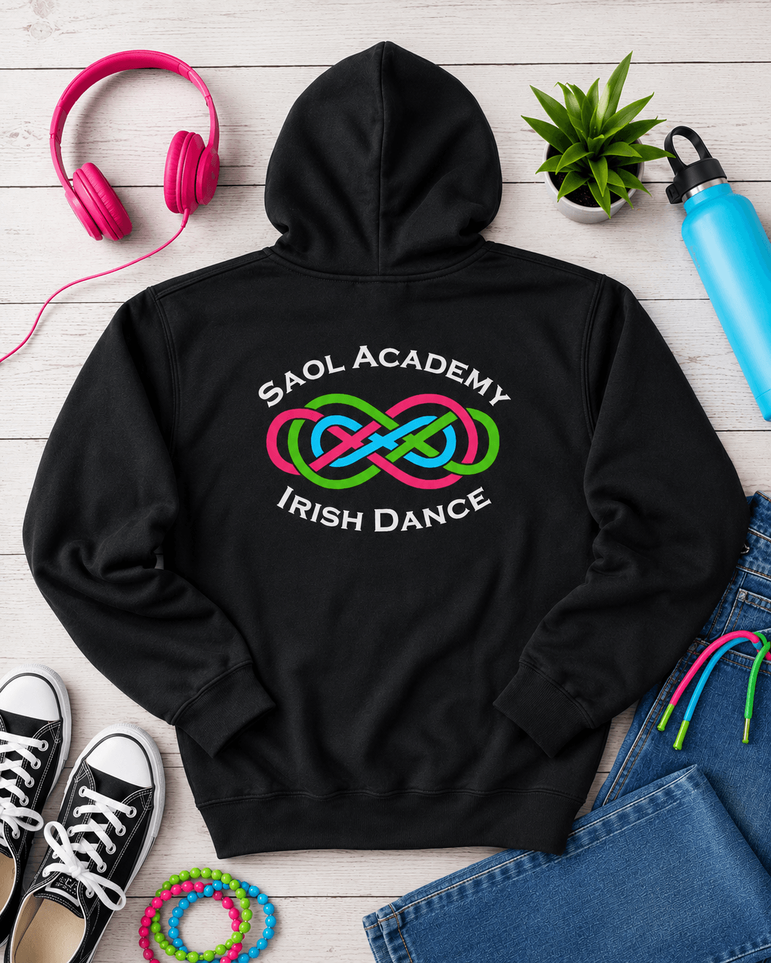 A black hoodie laid flat on a light wood surface, featuring a colorful graphic on the back that reads 'SAOL ACADEMY IRISH DANCE' around a pink, green, and blue infinity knot. Surrounding props include pink headphones, a small green plant, a blue water bottle, black sneakers, jeans, and multicolored wristbands.