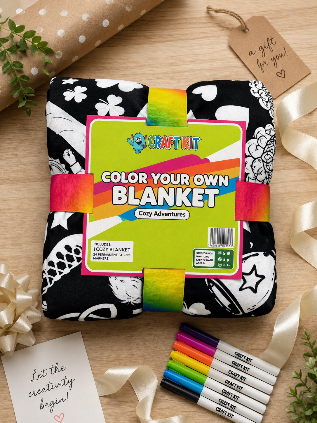 A packaged craft kit showing a folded black-and-white patterned blanket with a bright green label that reads 'COLOR YOUR OWN BLANKET' and 'Cosy Adventures.' The kit is styled on a wooden surface with ribbon, markers, wrapping paper, and small cards.