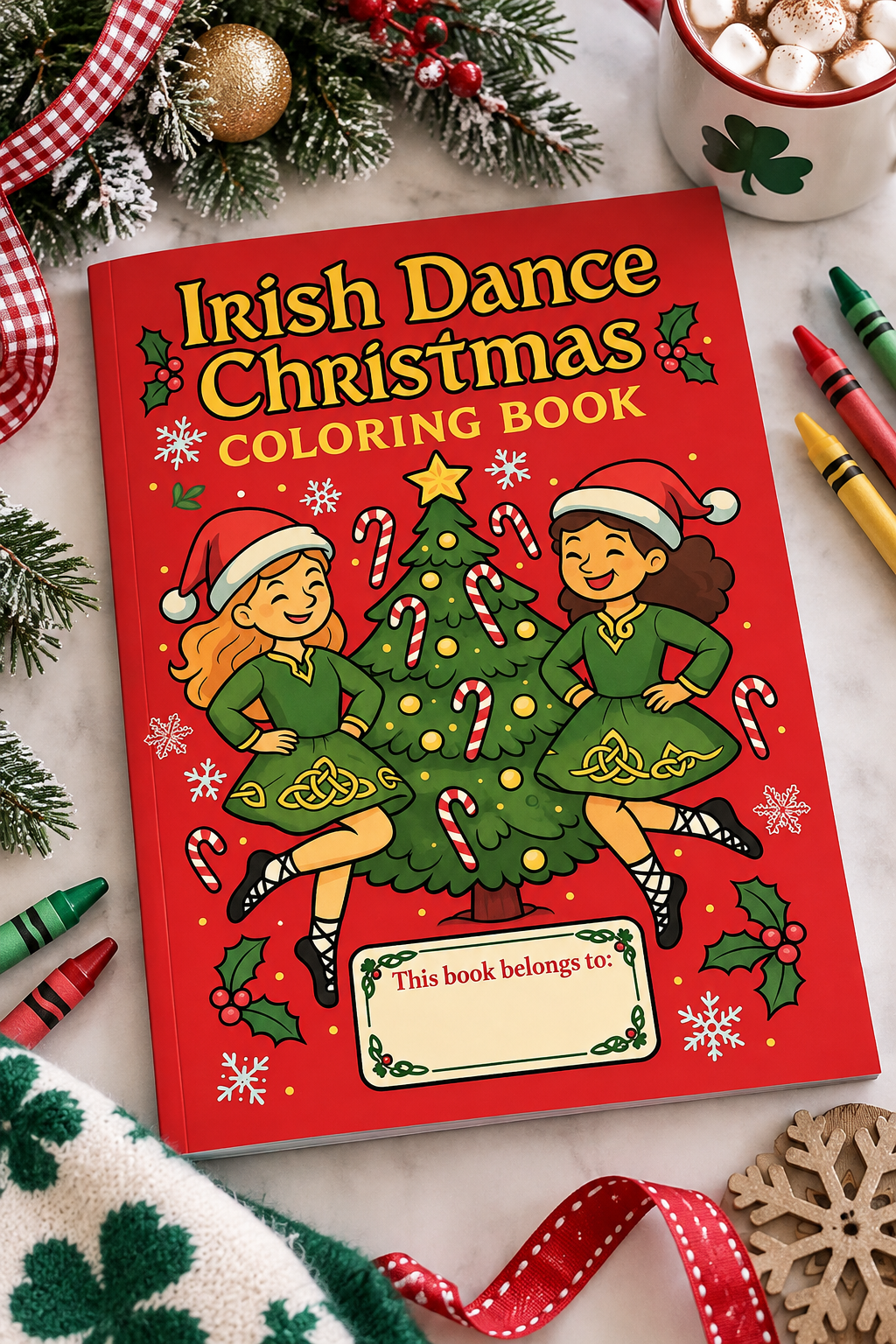A red hardcover coloring book titled 'Irish Dance Christmas Coloring Book' is shown in a styled flat-lay with Christmas decorations, crayons, a mug of marshmallows, and a patterned fabric. The cover features cartoon illustrations of two children dancing around a decorated Christmas tree.