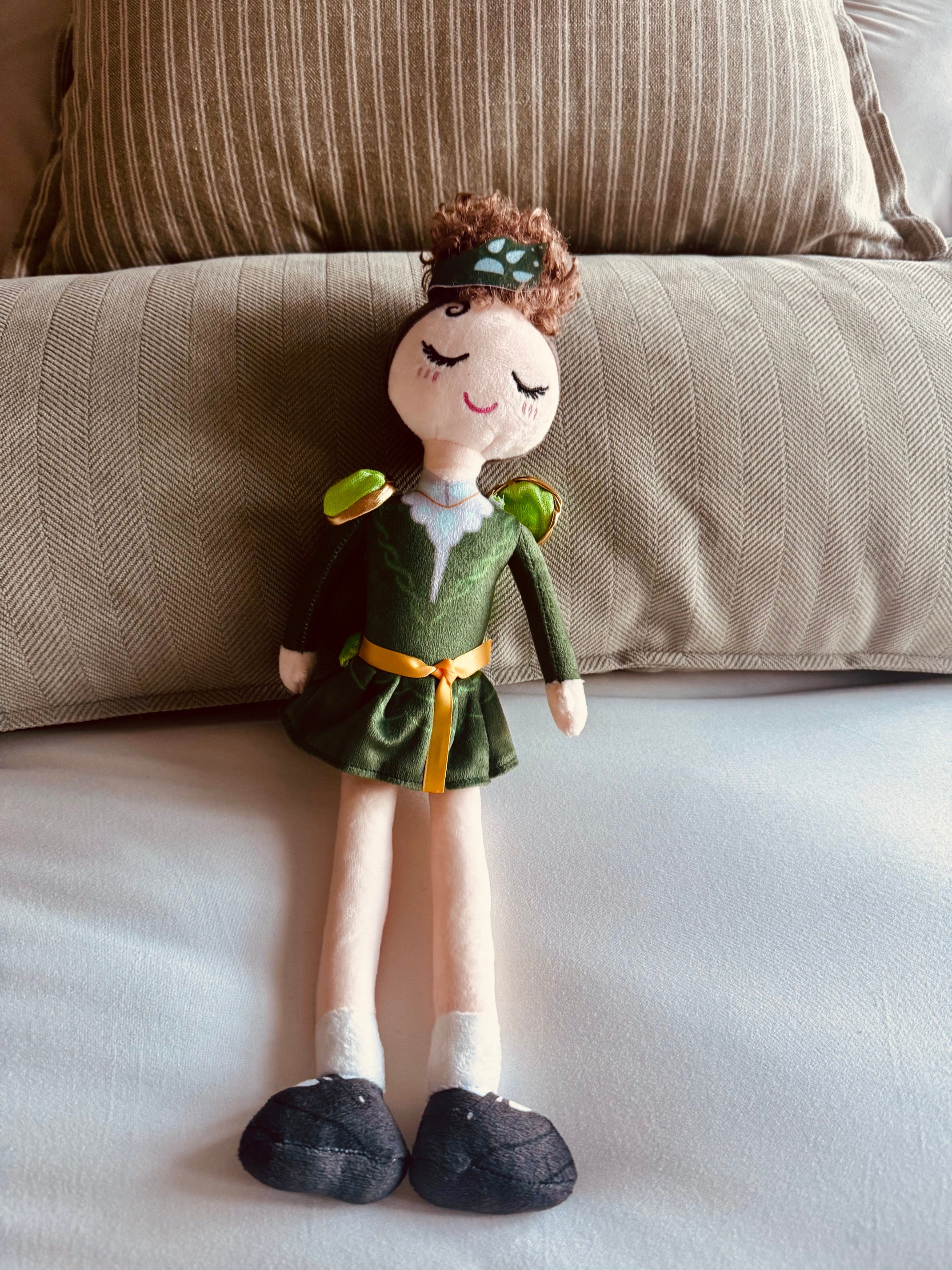 A handmade plush doll with brown curly hair, wearing a green dress with a yellow sash and green shoulder accents, posed on a white surface in front of a beige textured pillow.