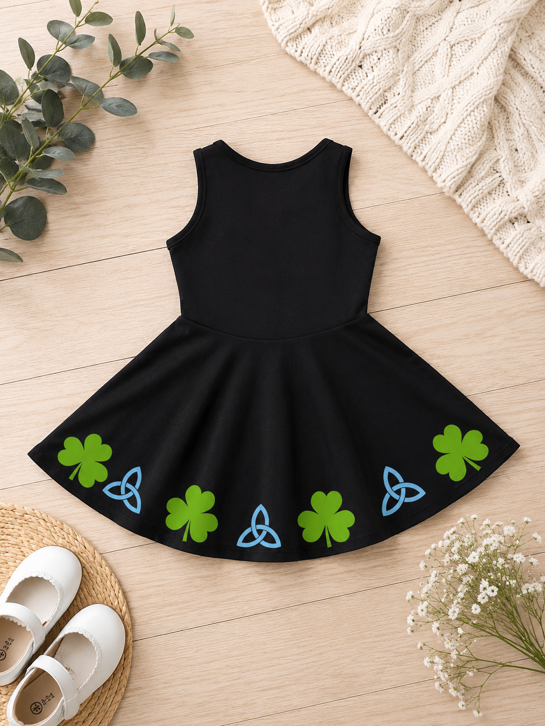 A sleeveless black dress with a flared skirt decorated with green shamrocks and blue triquetras, styled flat-lay on a light wood background with white shoes, eucalyptus, baby's breath, and a cream knit blanket.