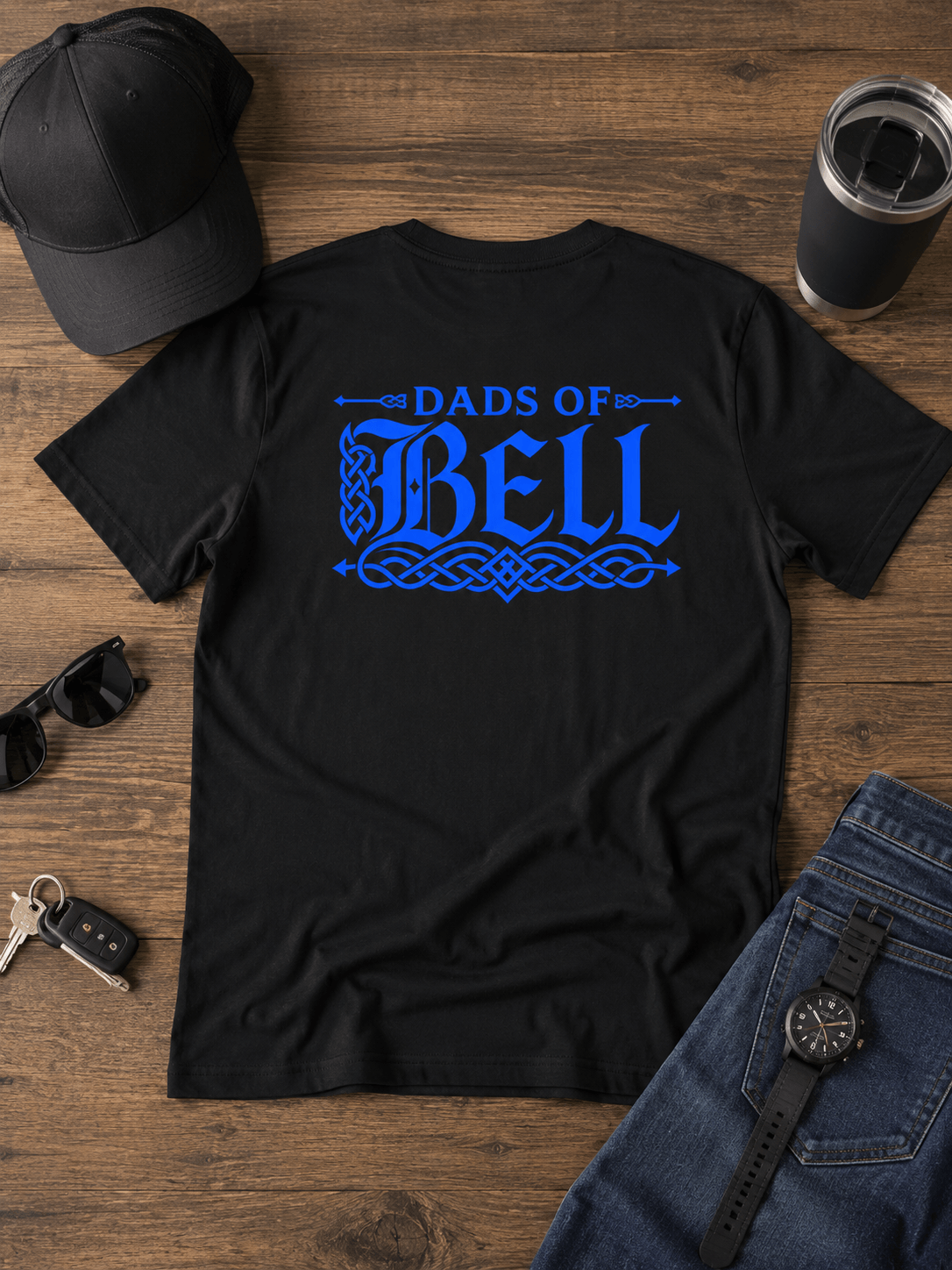 A black short-sleeve T-shirt laid flat on a wooden surface, featuring a large blue graphic text reading 'Dads of Bell' across the chest. The shirt is styled with a black baseball cap, sunglasses, keys, a tumbler, and blue jeans around it.