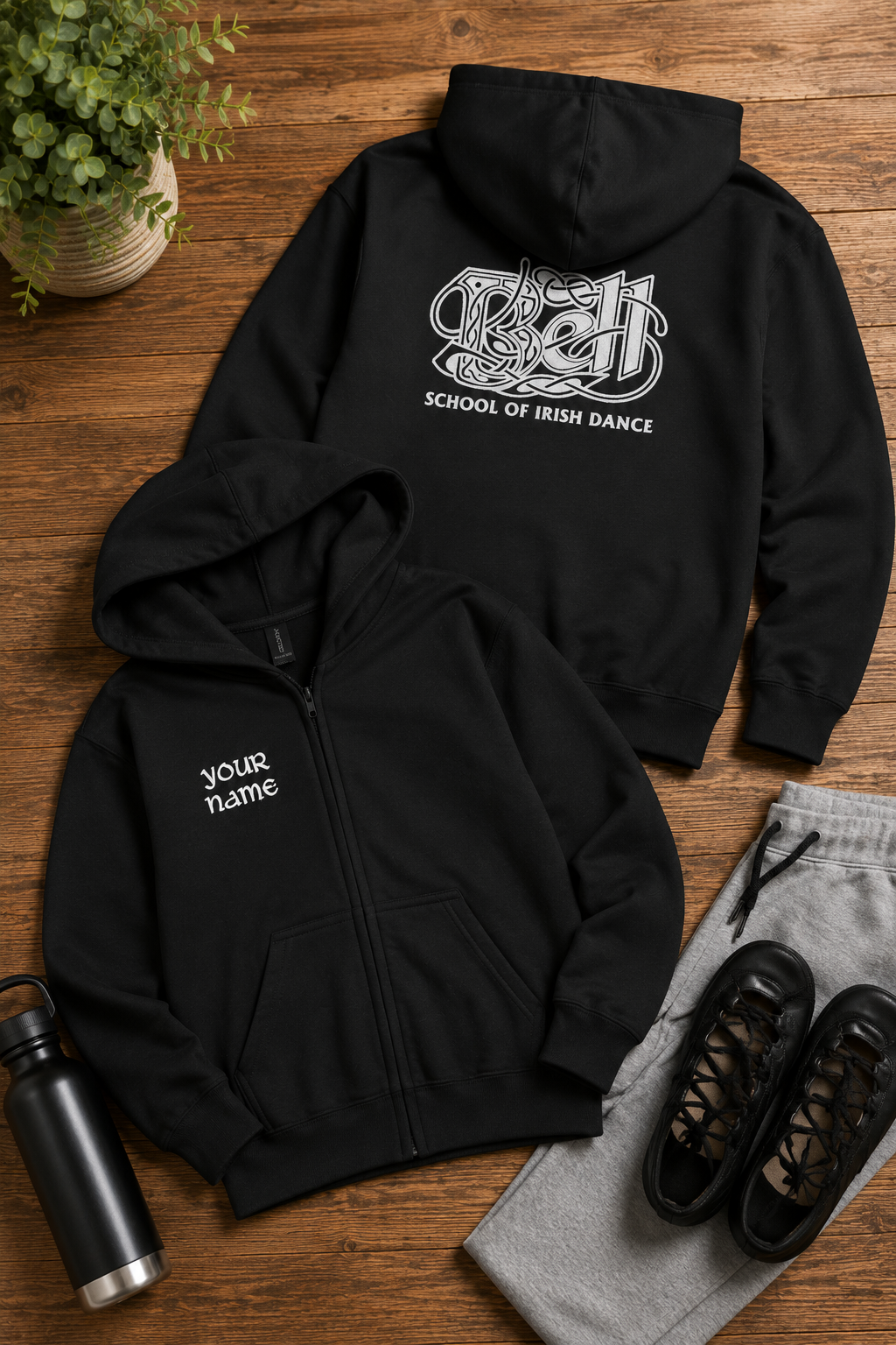 Two black zip-up hoodies laid flat on a wooden surface, one showing the front with 'Your name' text and the other showing the back with a white Celtic knot graphic and 'SCHOOL OF IRISH DANCE' text. Styled with gray sweatpants, black sneakers, a black water bottle, and a potted plant.