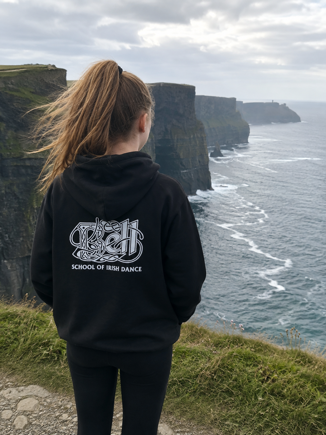 A person standing on a grassy cliff overlooking the Cliffs of Moher and ocean, wearing a black hoodie with a white graphic that reads 'School of Irish Dance' on the back.