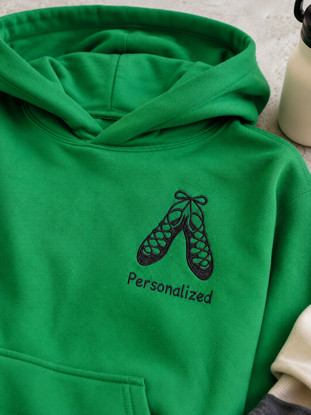 A bright green hoodie laid flat, featuring a black embroidered graphic of Irish dance shoes with the word 'Personalized' underneath. A beige sleeve is visible on the right edge.