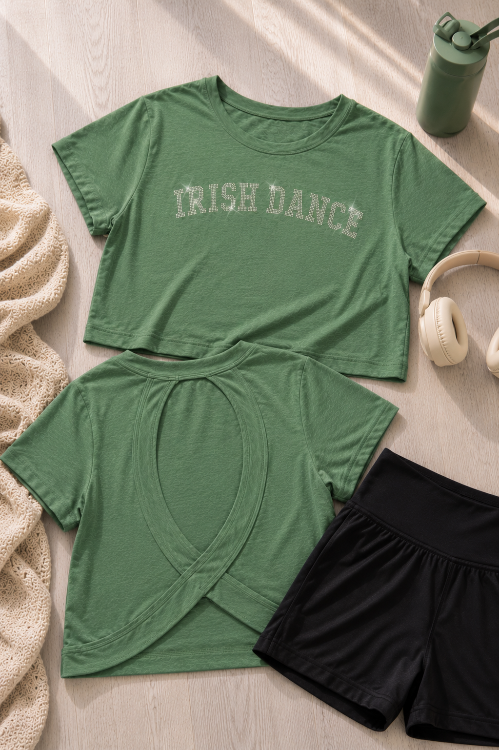 Two green short-sleeve crop top shirts shown flat-laid on a light wood floor, one with 'IRISH DANCE' text across the chest and one showing the back design. Black athletic shorts, a green tumbler, cream headphones, and a knit blanket are arranged around them as styling props.